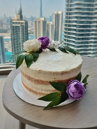 Naked vanilla cake with purple and white flowers by a window with the Dubai skyline